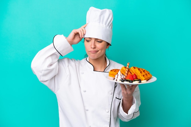 Young caucasian chef woman holding waffles isolated on blue background having doubts and with confuse face expression