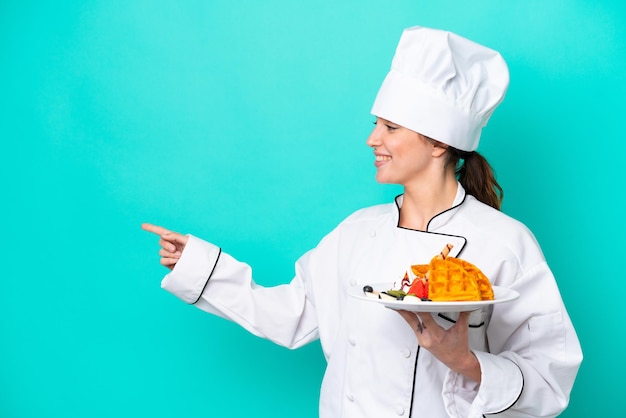 Young caucasian chef woman holding waffles isolated on blue background pointing to the side to present a product