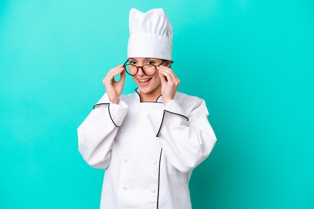 Young caucasian chef woman isolated on blue background with glasses and surprised