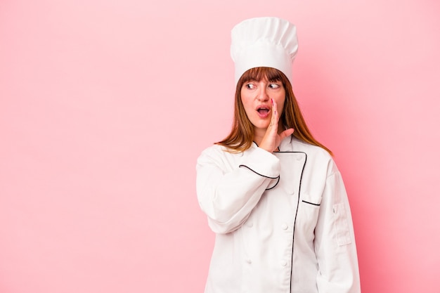 Young caucasian chef woman isolated on pink background is saying a secret hot braking news and looking aside