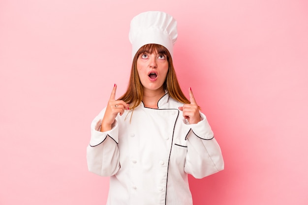 Young caucasian chef woman isolated on pink background pointing upside with opened mouth.