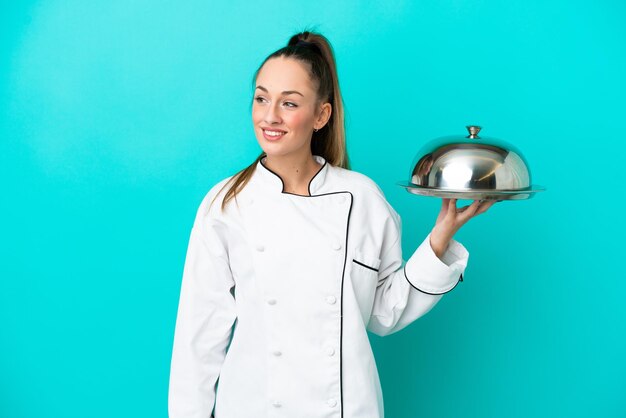 Молодая кавказская женщина-повар с подносом на синем фоне смотрит в сторону и улыбается