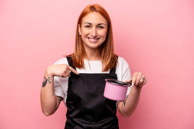 Young caucasian cooker woman holding translator isolated on pink background person pointing by hand to a shirt copy space, proud and confident