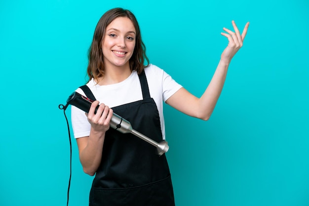 Young caucasian cooker woman using hand blender isolated on blue background extending hands to the side for inviting to come