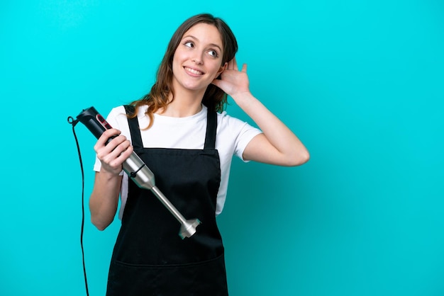 Young caucasian cooker woman using hand blender isolated on blue background listening to something by putting hand on the ear