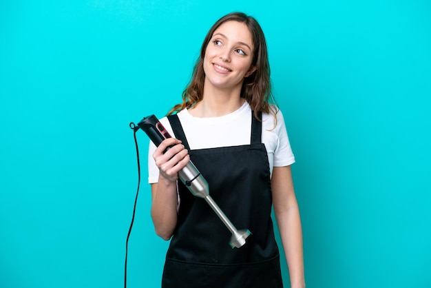 Young caucasian cooker woman using hand blender isolated on blue background thinking an idea while looking up