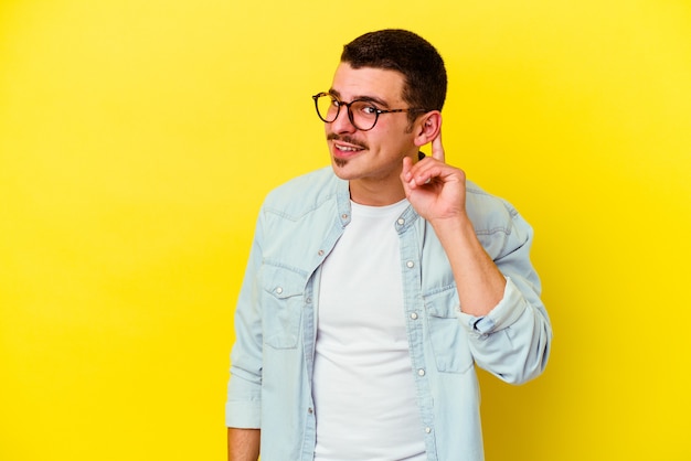 Young caucasian cool man isolated on yellow trying to listening a gossip.