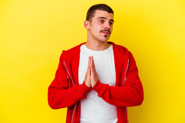 Young caucasian cool man isolated on yellow wall praying, showing devotion, religious person looking for divine inspiration