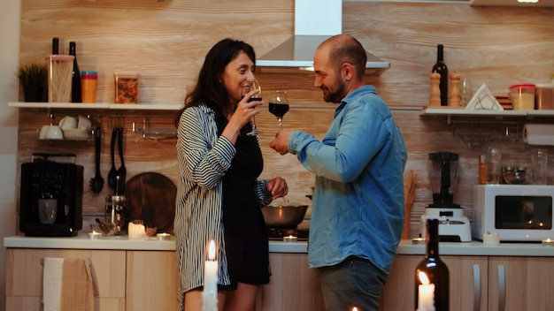 Young caucasian couple having romantic date at home in the kitchen, drinking red wine, talking, smiling in dining room. Two people in love having pleasant conversation during healthy meal.