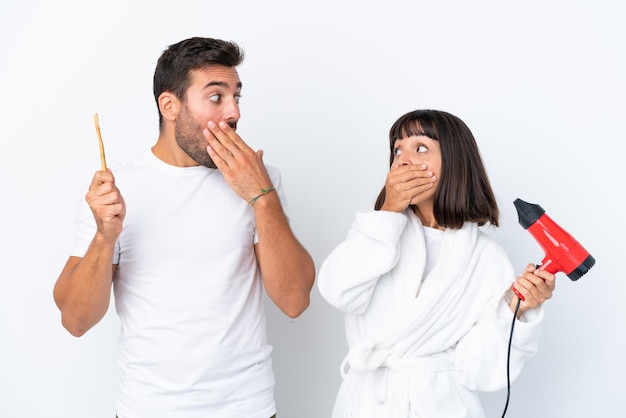 Young caucasian couple holding a hairdryer and toothbrush isolated on white background covering mouth with hands for saying something inappropriate