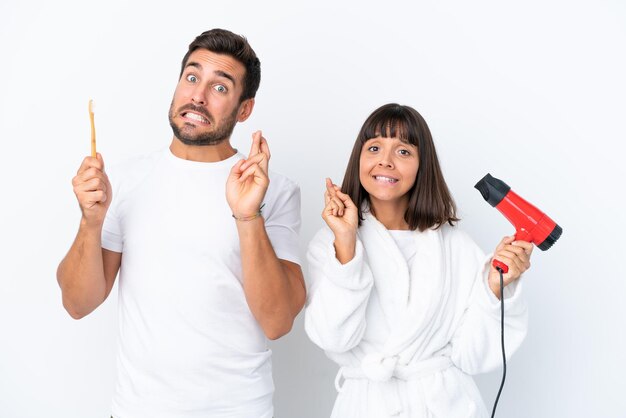 Young caucasian couple holding a hairdryer and toothbrush isolated on white background with fingers crossing and wishing the best