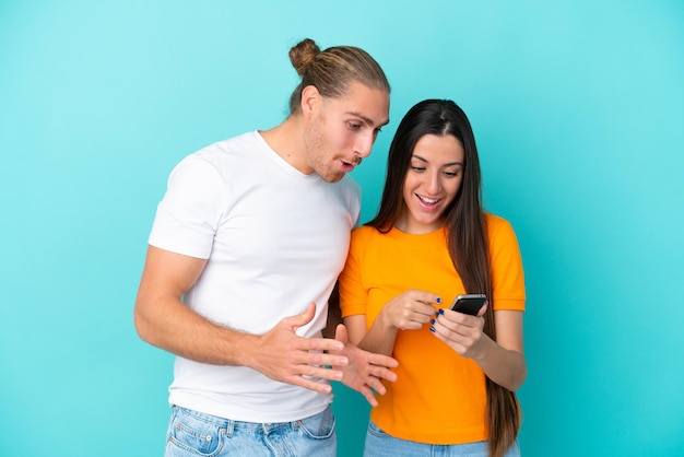 Young caucasian couple isolated on blue background sending a message or email with the mobile