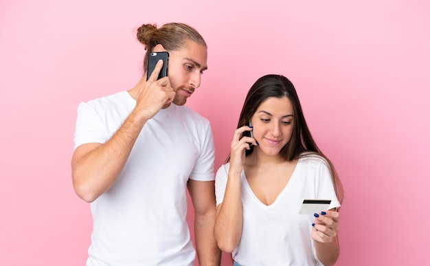 Young caucasian couple isolated on pink background buying with the mobile with a credit card