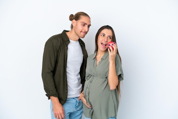 Young caucasian couple isolated on white background pregnant and holding an apple and eating it