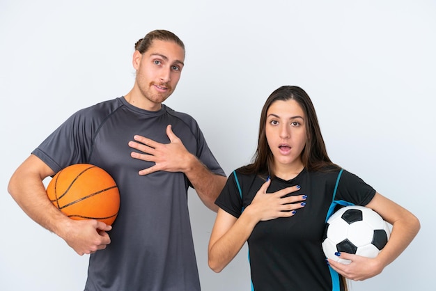 Young caucasian couple playing basketball and football isolated on white background surprised and shocked while looking right