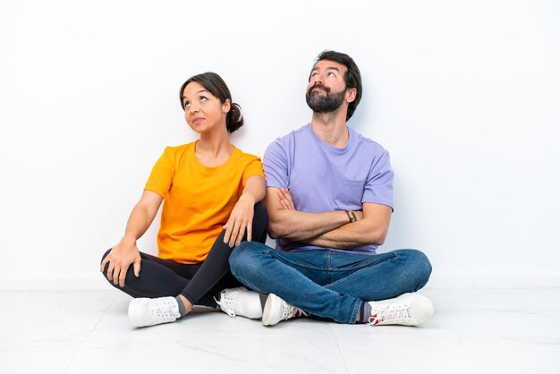 Photo young caucasian couple sitting on the floor isolated on white background looking up while smiling