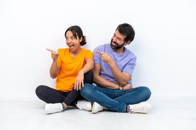 Young caucasian couple sitting on the floor isolated on white background pointing finger to the side