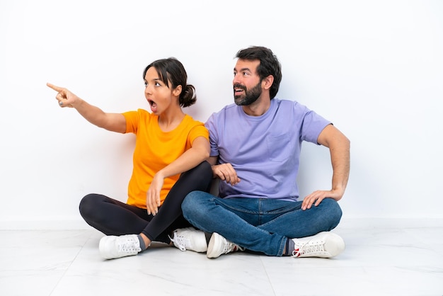 Young caucasian couple sitting on the floor isolated on white background presenting an idea while looking smiling towards