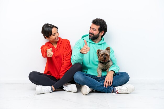 Young caucasian couple sitting on the floor with their pet isolated on white background giving a thumbs up gesture