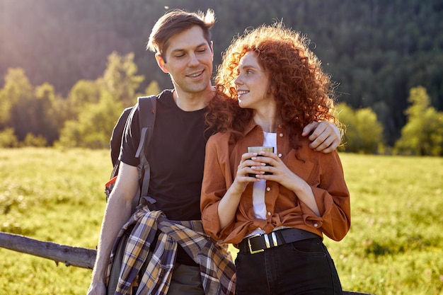 Young caucasian couple spending time in nature. Visiting countryside,active tourism. Hiking in the mountains vacation concept, have rest, drinking tea from thermos.Peaceful rural environment.