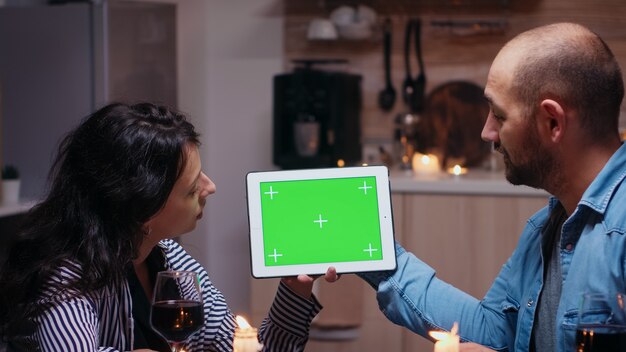Молодая кавказская пара с помощью цифрового изолированного планшетного компьютера зеленого экрана макета. Муж и жена, глядя на экран хроматического ключа шаблона зеленый экран, сидя за столом на кухне во время обеда.