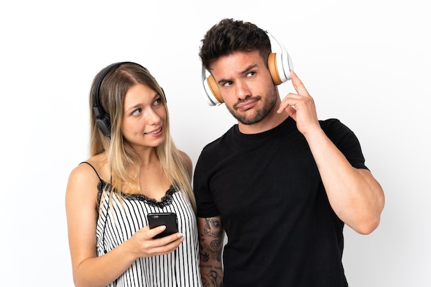 Young caucasian couple on white listening music with a mobile and thinking