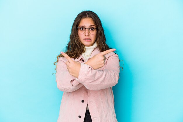 Young caucasian curly hair woman isolated points sideways, is trying to choose between two options.