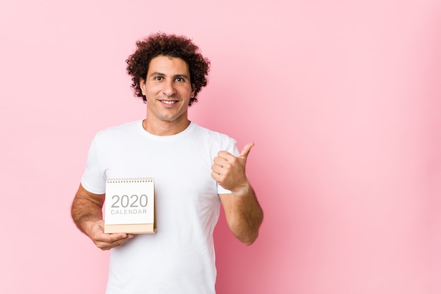 Young caucasian curly man holding a 2020 calendar smiling and raising thumb up