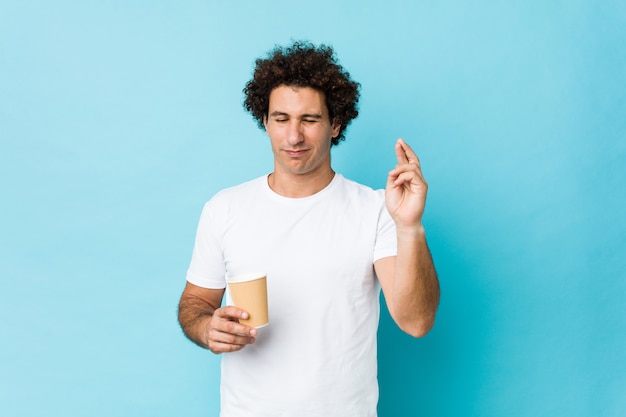 Young caucasian curly man holding a takeaway coffee crossing fingers for having luck