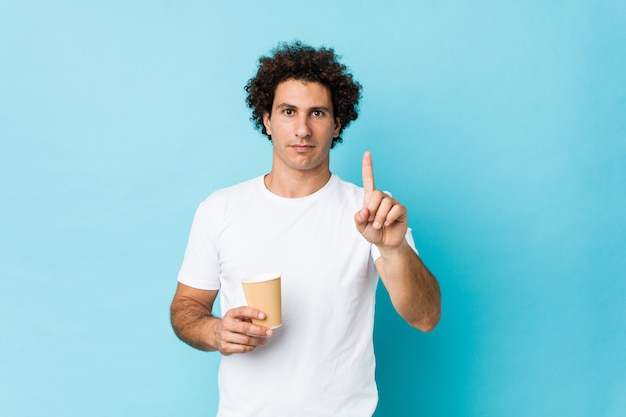 Young caucasian curly man holding a takeaway coffee showing number one with finger.