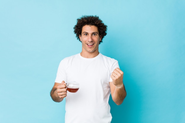 Young caucasian curly man holding a tea cup cheering carefree and excited