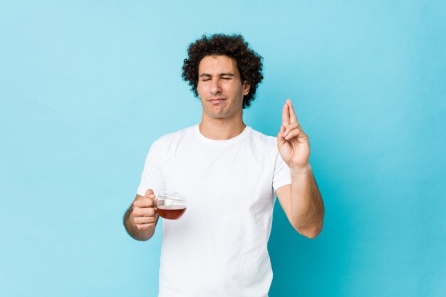 Young caucasian curly man holding a tea cup crossing fingers for having luck