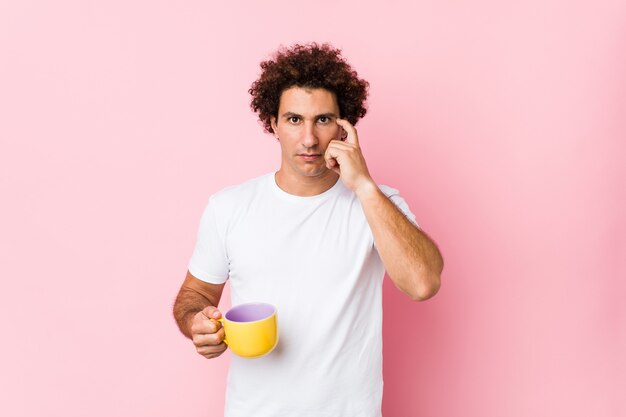 Young caucasian curly man holding a tea cup pointing his temple with finger, thinking, focused on a task.