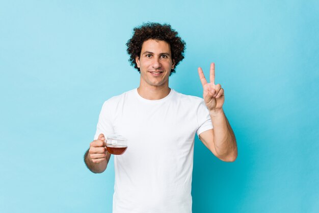 Young caucasian curly man holding a tea cup showing victory sign and smiling broadly.