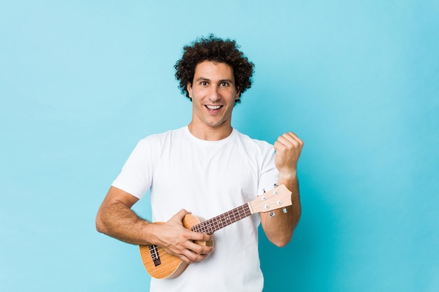 Young caucasian curly man playing ukelele cheering carefree and excited. Victory concept.