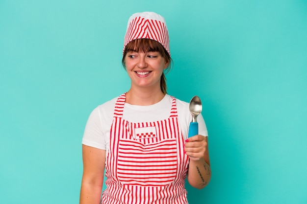 Молодой кавказский пышный производитель мороженого, держащий совок, изолированный на синем фоне, смотрит в сторону улыбающимся, веселым и приятным.