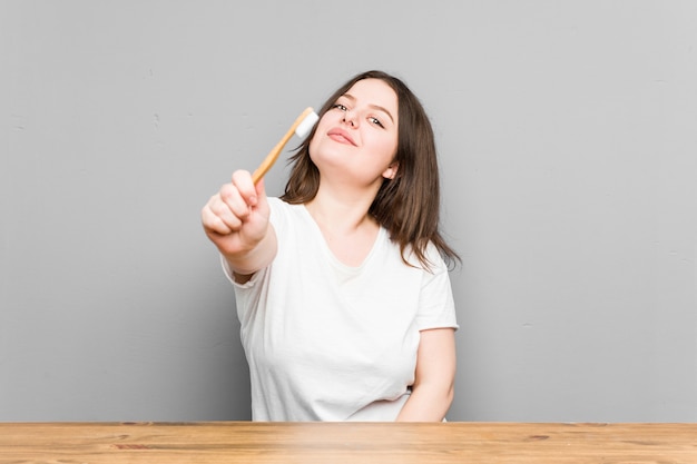 Giovane donna curvy caucasica che pulisce i suoi denti con uno spazzolino da denti isolato su una parete grigia