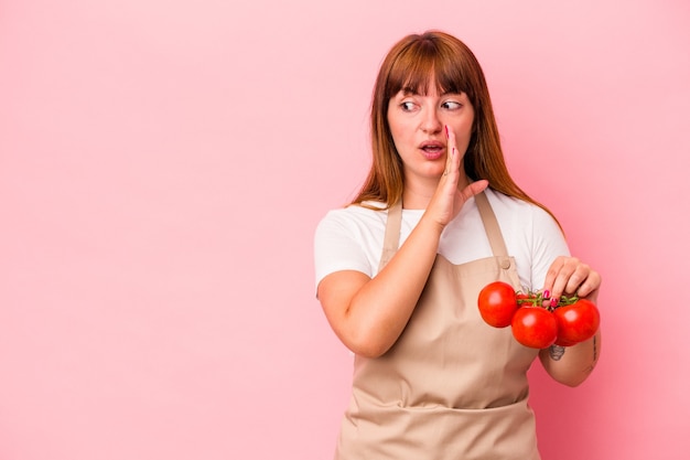 ピンクの背景に分離されたトマトを持って家で料理をしている若い白人の曲がりくねった女性は、秘密の熱いブレーキングニュースを言って、脇を見ています