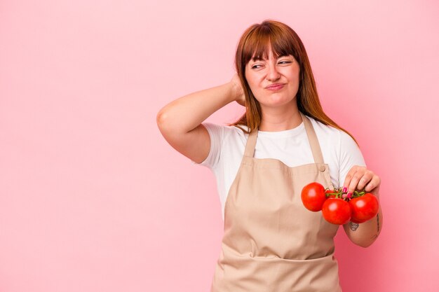 Young caucasian curvy woman cooking at home holding tomatoes isolated on pink background touching back of head, thinking and making a choice.