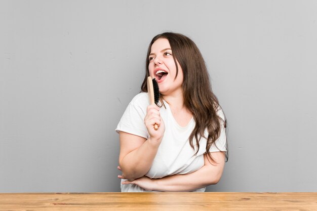 Young caucasian curvy woman holding a hair brush isolated on grey