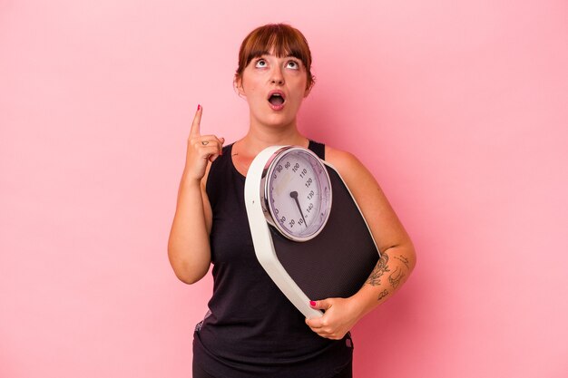 Young caucasian curvy woman holding weighing machine isolated on pink background pointing upside with opened mouth.