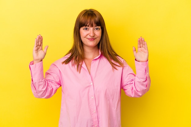 Young caucasian curvy woman isolated on yellow background holding something little with forefingers, smiling and confident.