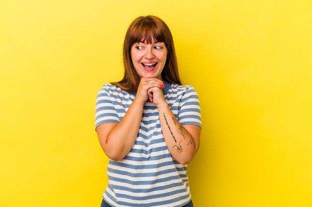 Young caucasian curvy woman isolated on yellow background keeps hands under chin, is looking happily aside.