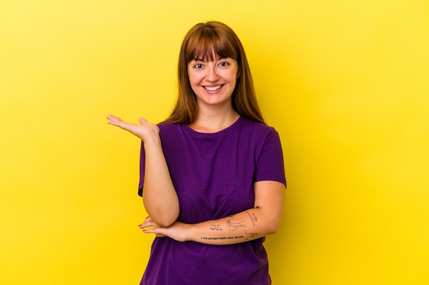 Young caucasian curvy woman isolated on yellow background showing a copy space on a palm and holding another hand on waist.