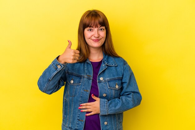 Young caucasian curvy woman isolated on yellow background touches tummy, smiles gently, eating and satisfaction concept.