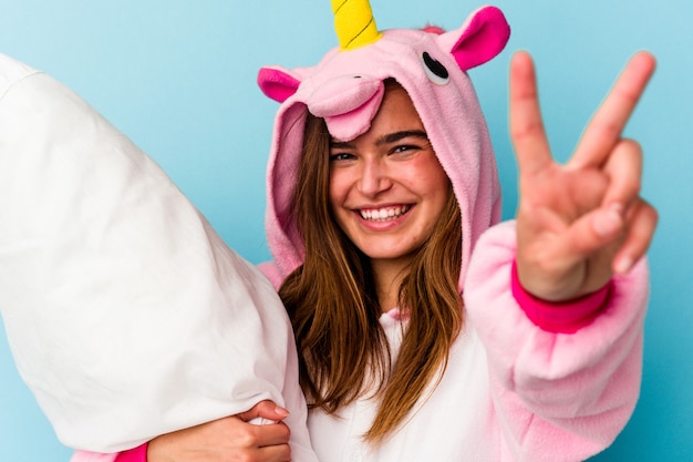 Young caucasian cute woman wearing an unicorn pajama holding a pillow isolated on blue
