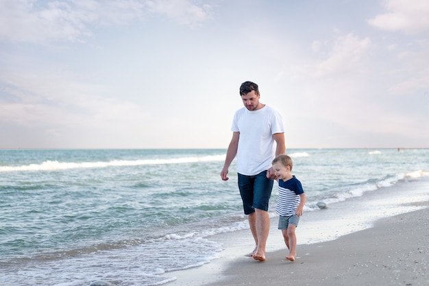 Young Caucasian dad with little son walk at sea beach warm summer day