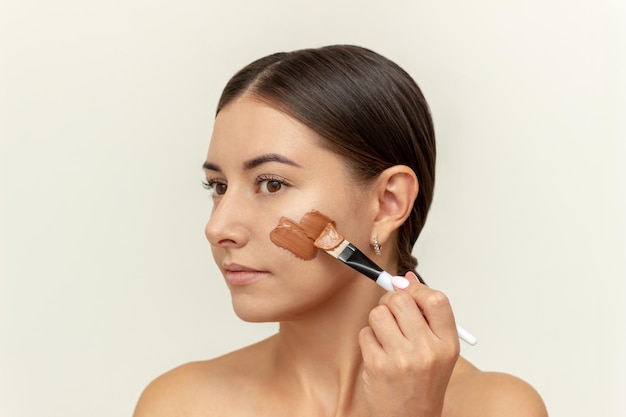 A young Caucasian dark-haired woman applies a red clay mask to her face with a brush