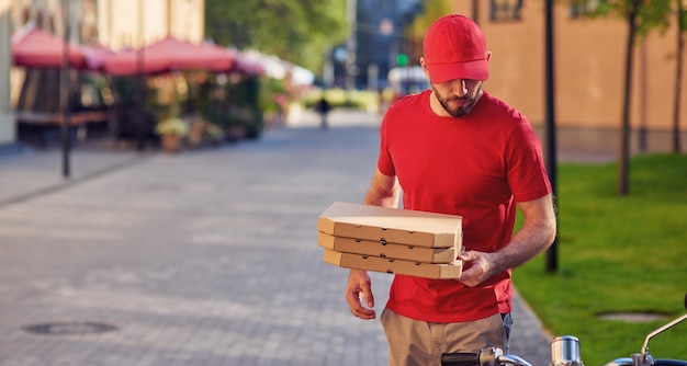 Young caucasian delivery man in red uniform delivering pizza, standing near his scooter, selective focus on courier. Food delivery concept