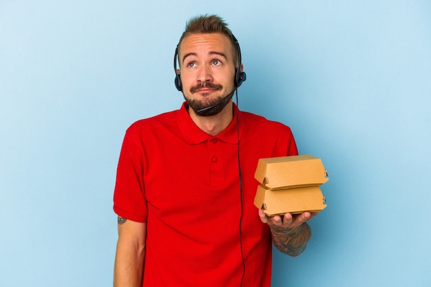 Young caucasian delivery man with tattoos holding burgers isolated on blue background  dreaming of achieving goals and purposes